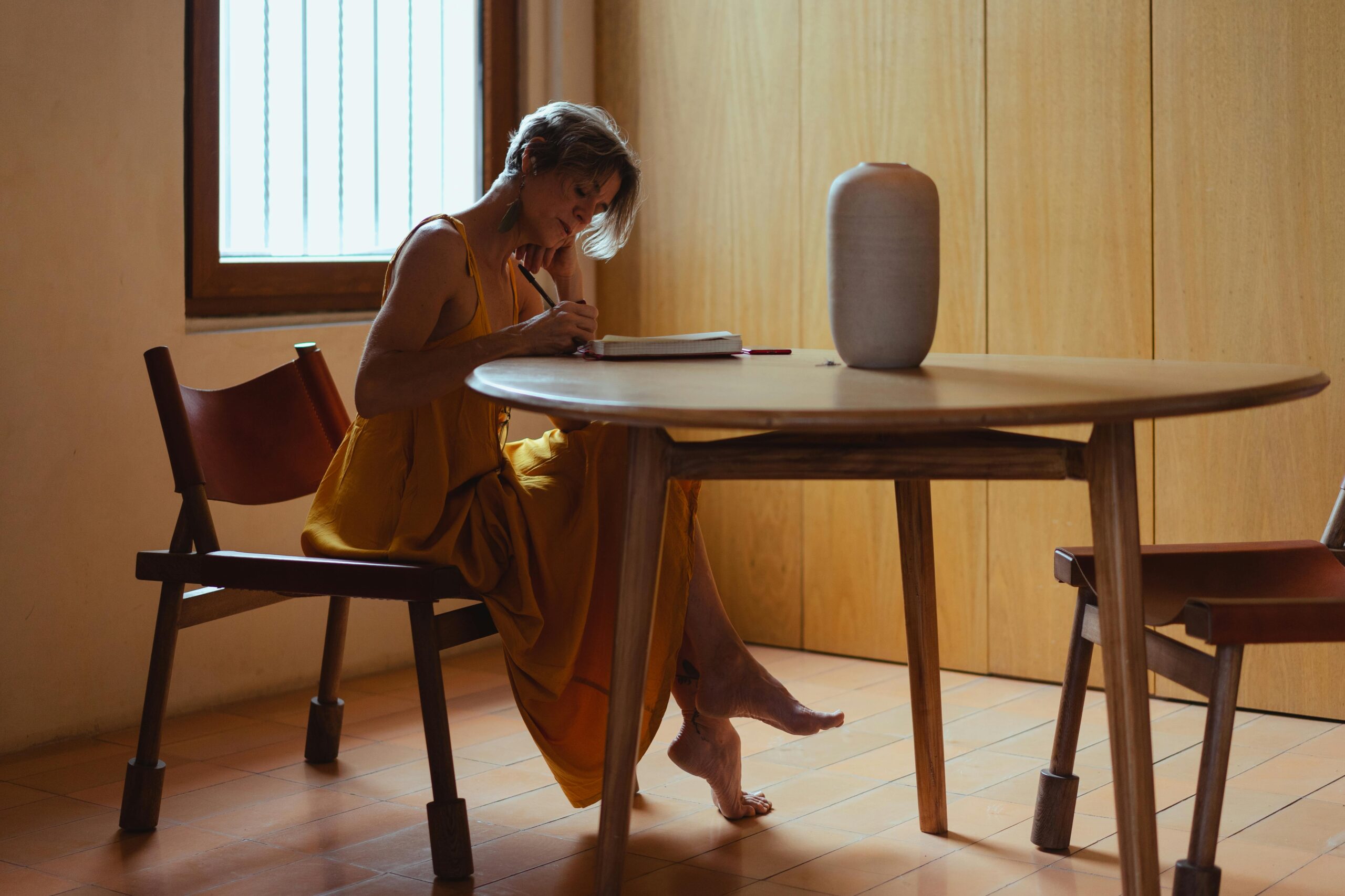 A senior woman in a yellow dress writes in a journal at a wooden table, creating a moment of reflection.